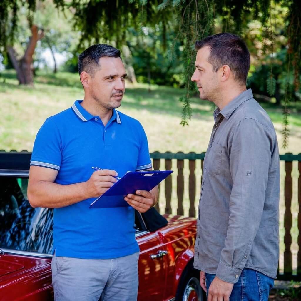 Zwei Männer unterhalten sich draußen neben einem roten Auto, einer hält ein Klemmbrett und prüft Reparaturfreigabe oder Versicherung
